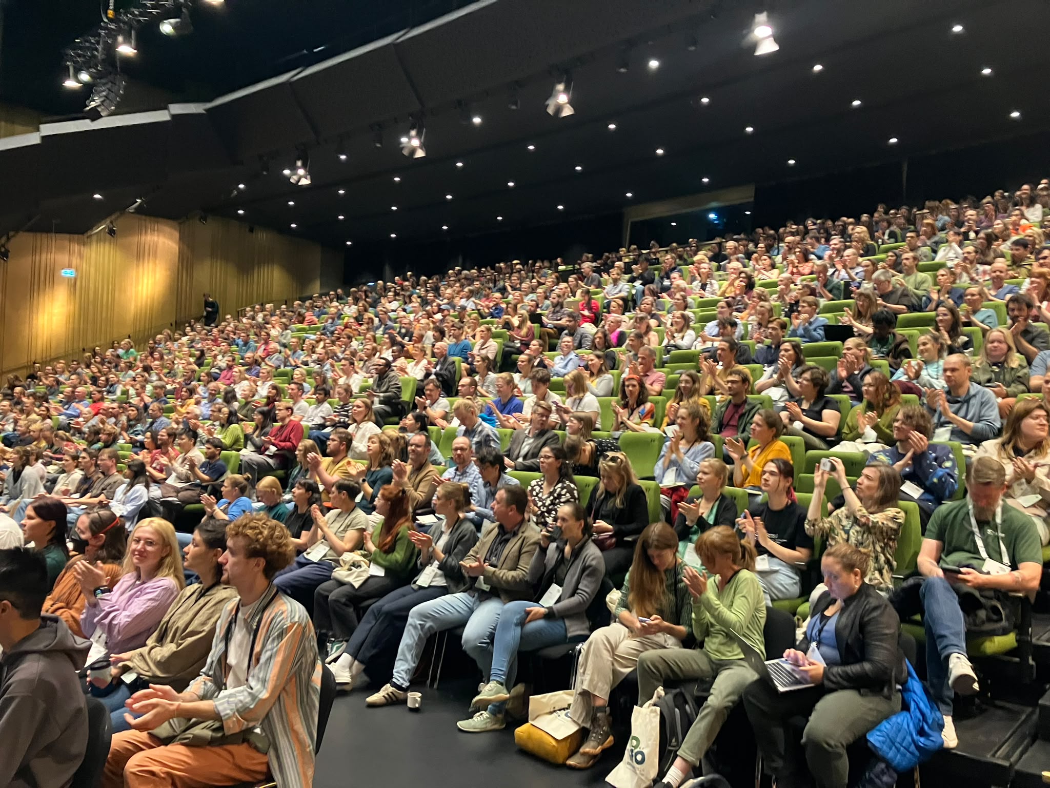 The image shows the inside of a large lecture theatre filled with people. 