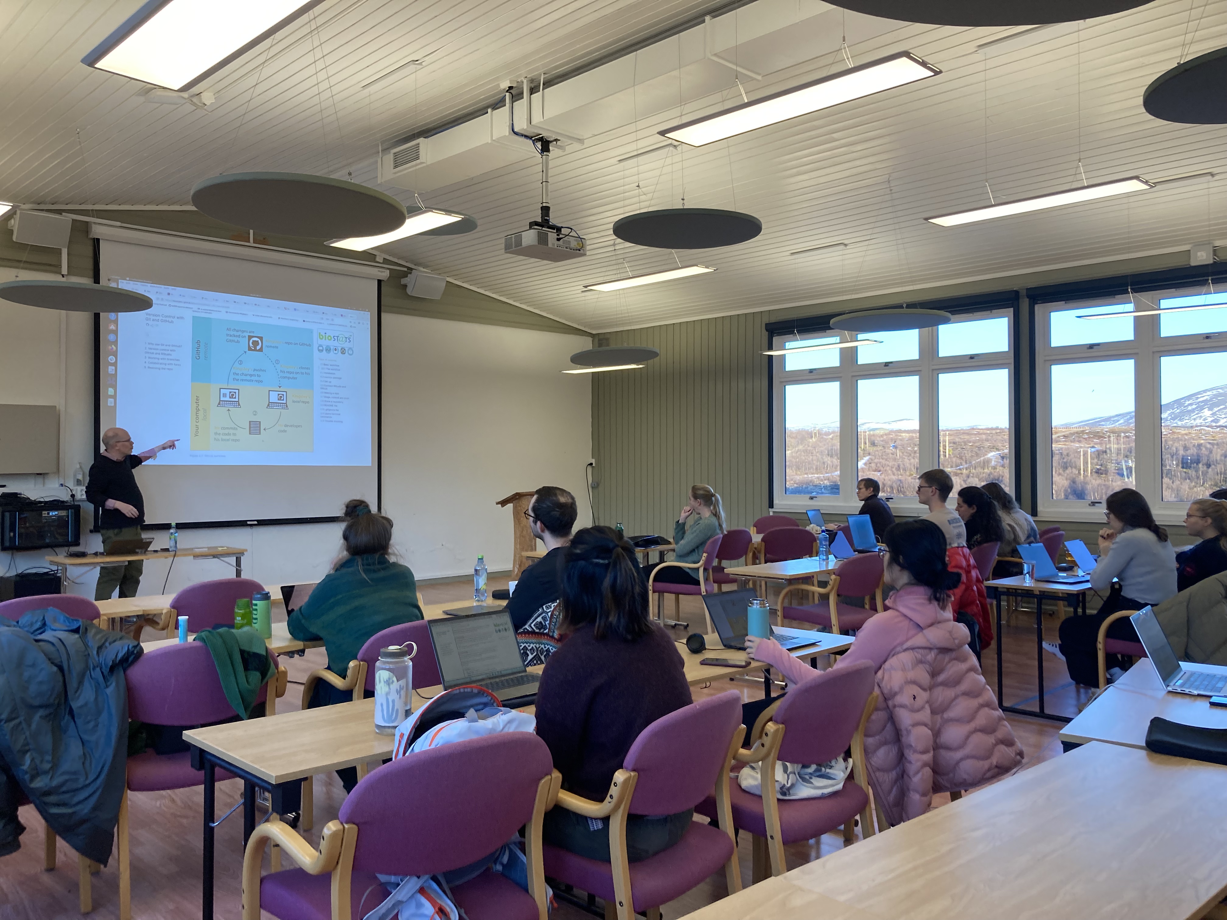 The image shows a seminar with a presenter at the front and several participants seated.